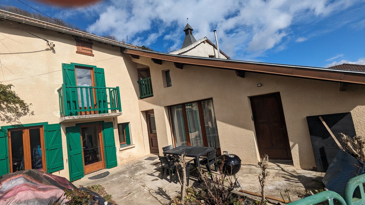 The exterior of the house features a light-colored facade with green shutters and a balcony. A spacious outdoor dining area is seen with a large table and chairs. Large windows invite natural light, while a nearby door provides access to the interior.
