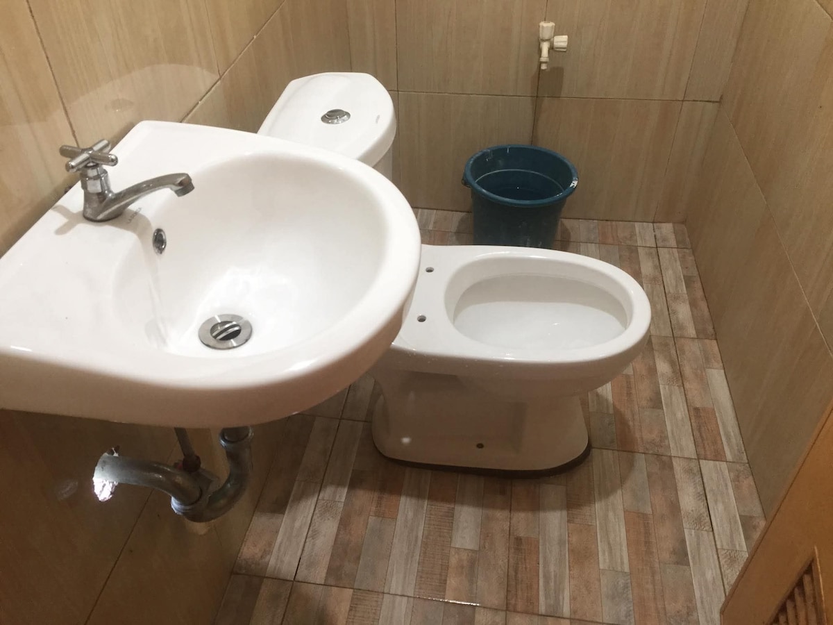 A compact bathroom features a sink with a chrome faucet and a modern toilet. Light-colored tiles cover the walls and floor, enhancing the clean and simple design. A blue bucket is positioned in the corner, adding a practical touch to the space.