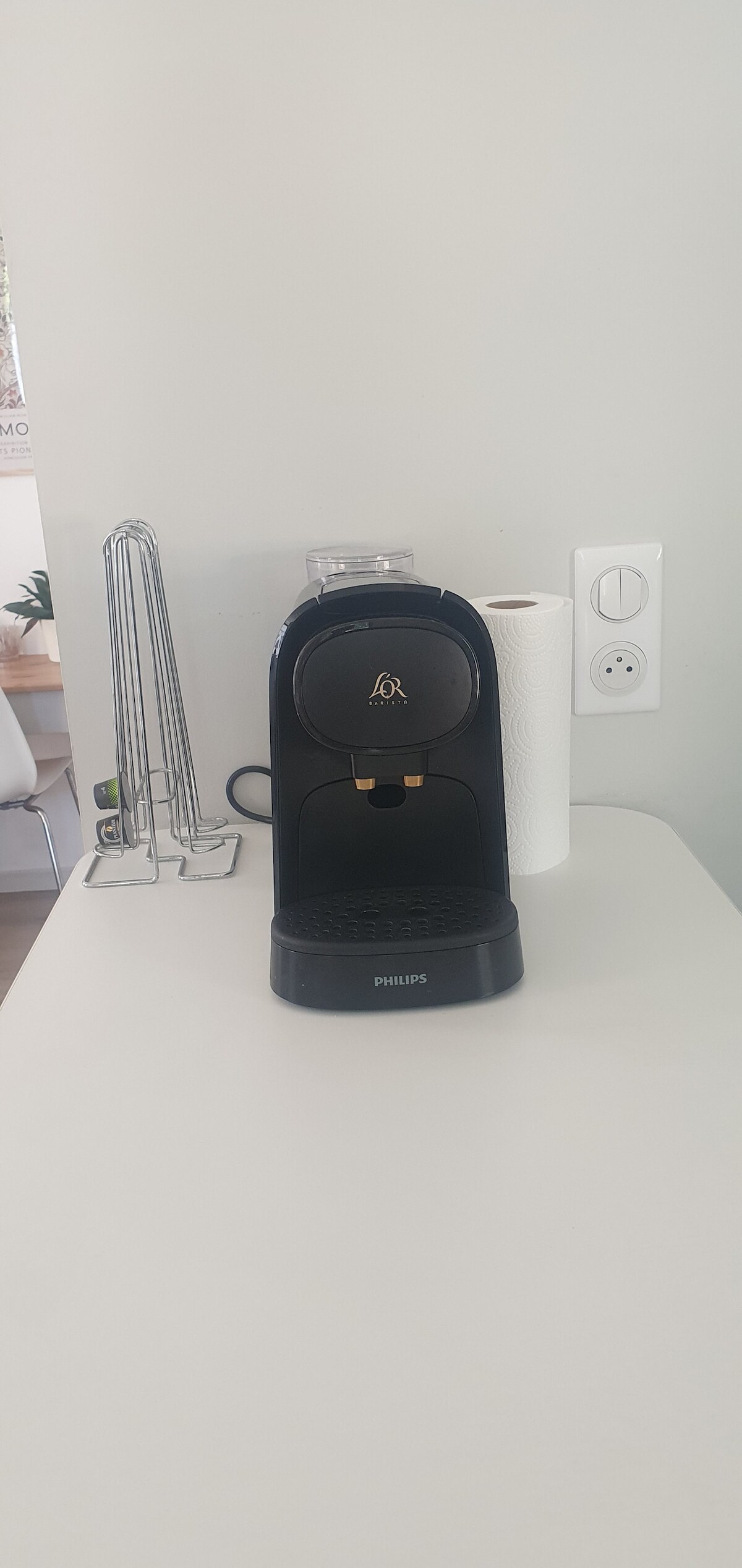 A coffee maker is positioned on a clean countertop, featuring a sleek black design. Nearby, a roll of paper towels and a tall utensil holder are visible, contributing to a functional kitchen space. Natural light enhances the bright and tidy environment.