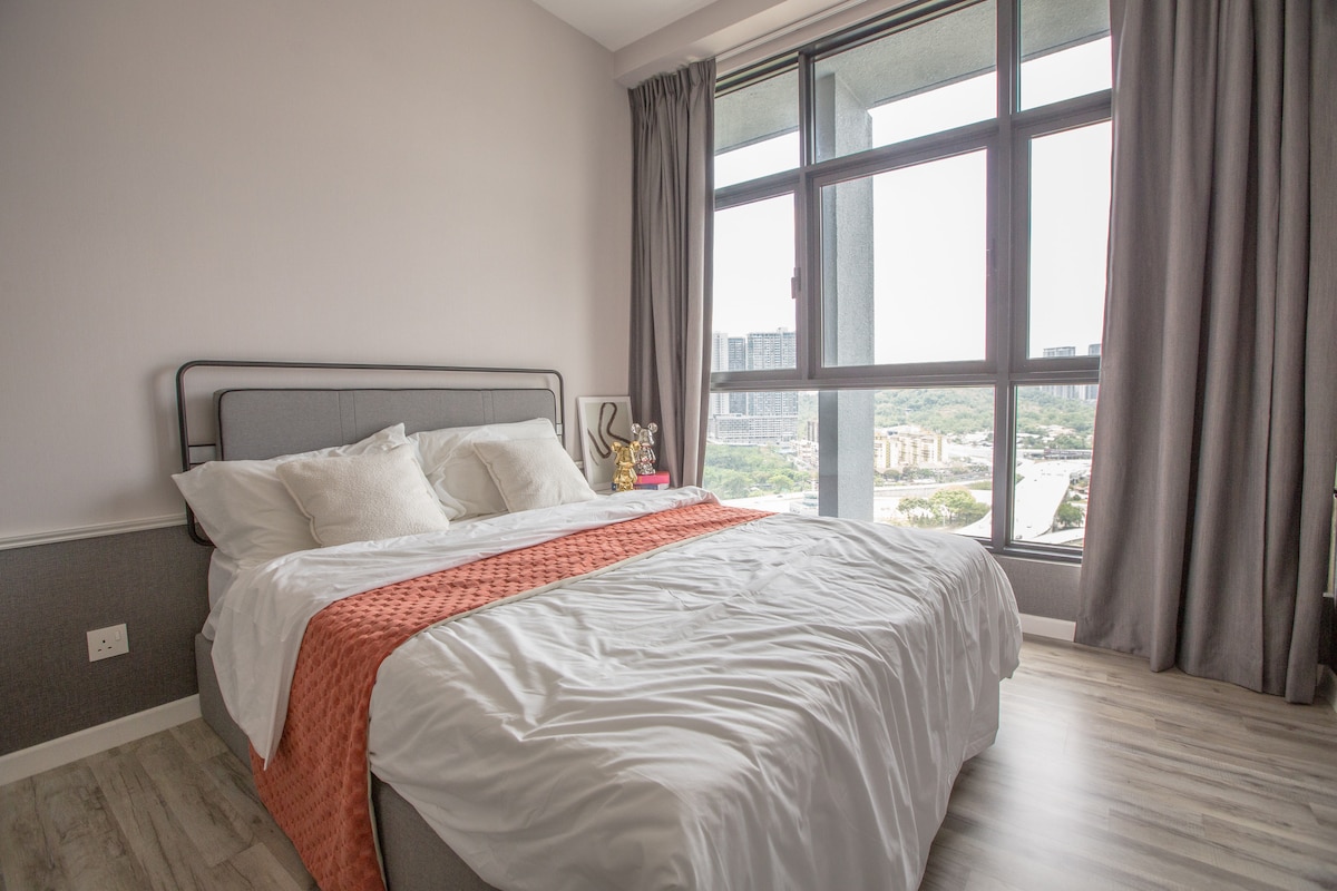 A bright bedroom features a spacious bed adorned with a coral-colored throw blanket. Large windows provide natural light and a view of the surrounding area, while gray curtains frame the windows, adding a subtle touch to the interior design.