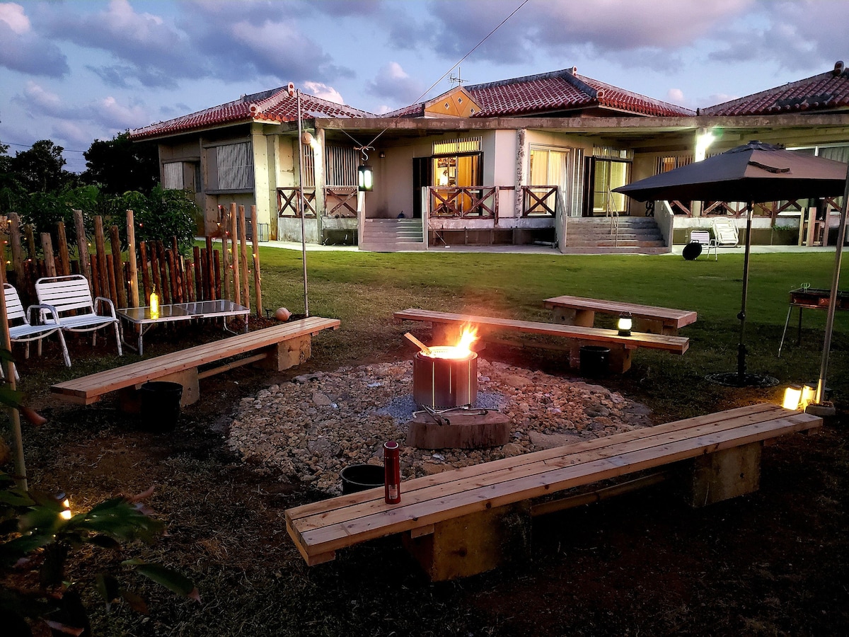 A cozy fire pit area is surrounded by wooden benches, creating a welcoming space for gathering. The backyard features soft grass, while nearby structures showcase traditional architecture, illuminated by warm lighting as the sun sets behind the clouds.