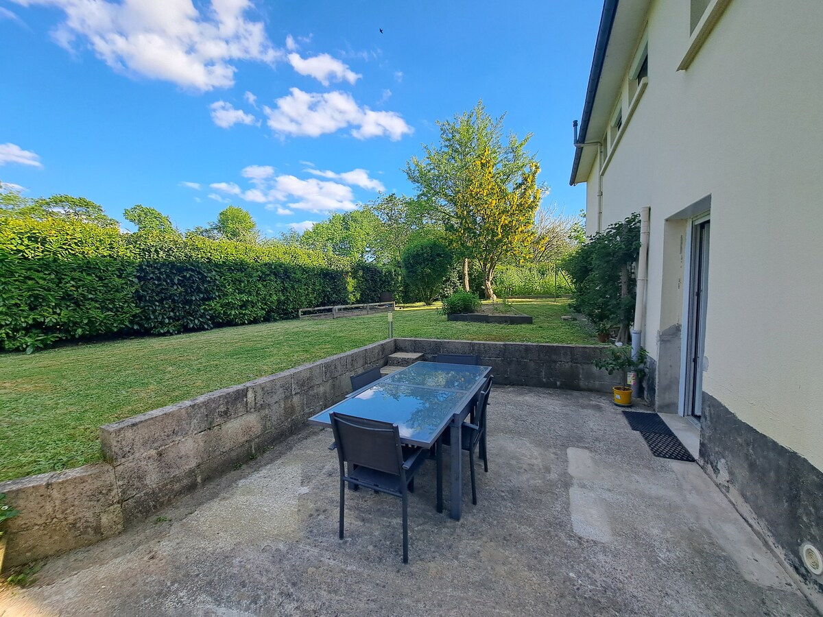 An outdoor patio features a glass table surrounded by four black chairs. The space overlooks a grassy yard enclosed by hedges and trees, under a bright blue sky with scattered clouds. The setting offers a peaceful environment for relaxation.