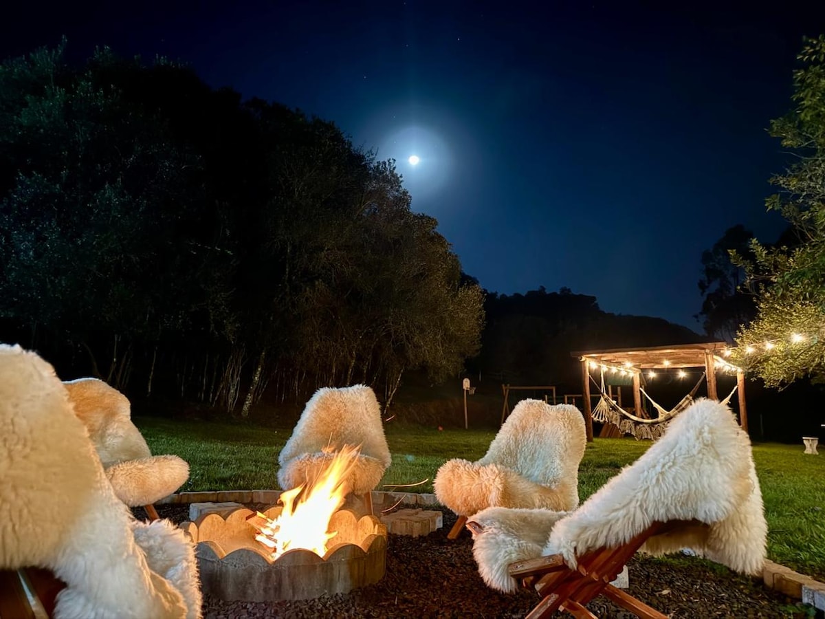 A fire pit is surrounded by cozy, fur-covered chairs, creating a warm gathering space. The night sky is illuminated by the moon, and a gazebo with string lights can be seen in the background among greenery.