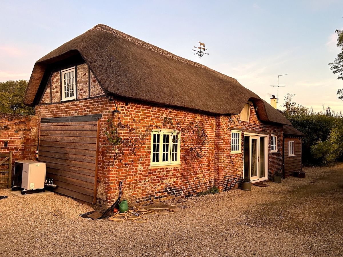 The exterior of a traditional coach house is showcased, featuring a charming thatched roof and red brick walls. Large windows and a set of glass doors provide a warm, inviting entrance, while the surrounding gravel area offers ample space for outdoor activities.