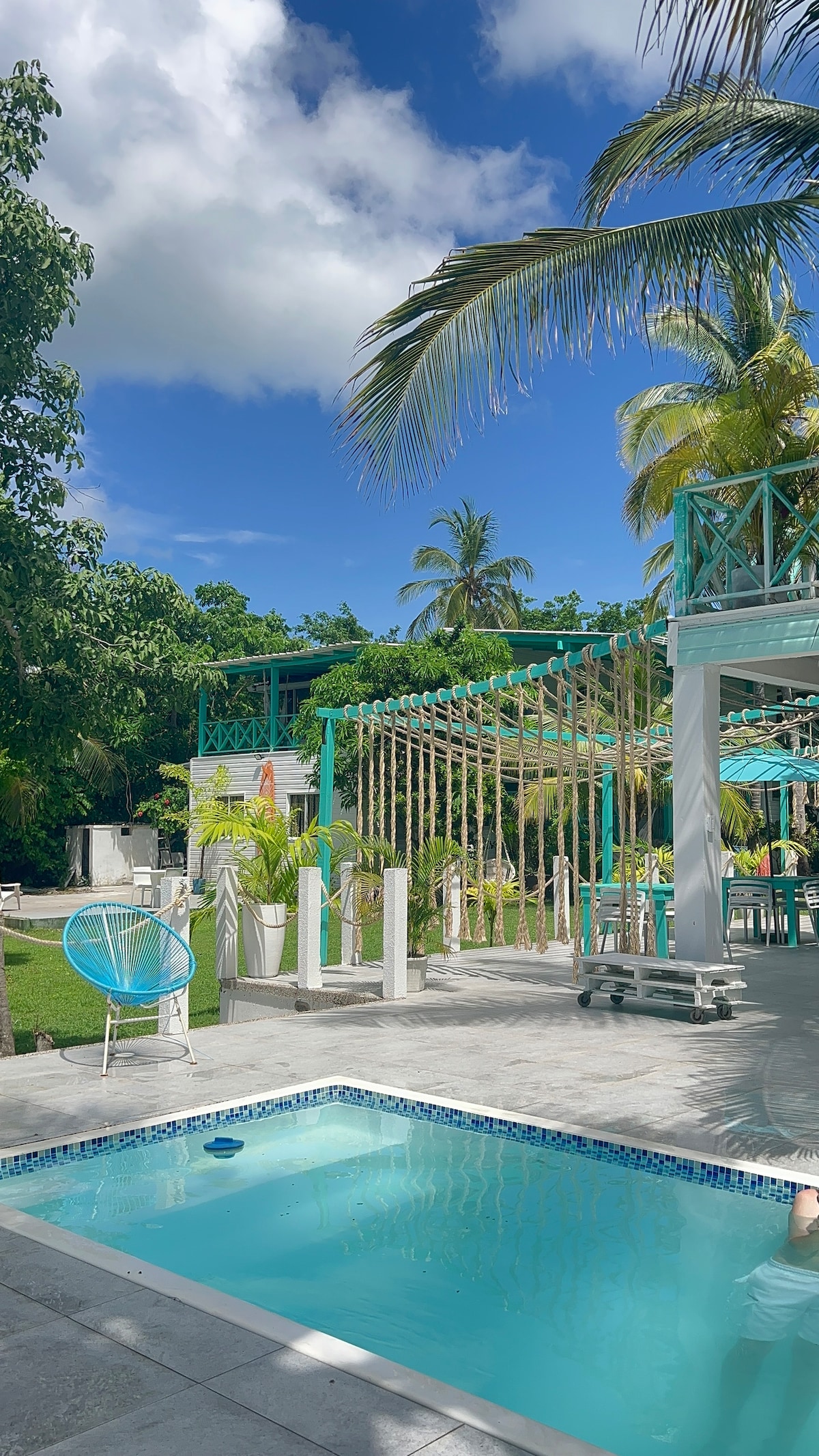 A serene pool area is surrounded by lush greenery, featuring a small, inviting swimming pool with clear blue water. Colorful lounge seating is positioned nearby under the shade of palm trees, creating a relaxing atmosphere for guests to enjoy.