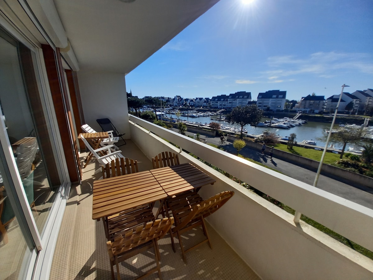 A balcony features a wooden dining table surrounded by folding chairs, offering views of the marina and surrounding residential area. Sunlight brightens the space, creating a suitable spot for outdoor meals. A sun lounger is positioned to the side, adding to the relaxing atmosphere.