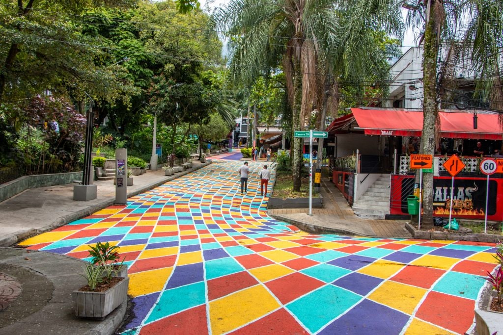 A vibrant street is illustrated with a colorful patterned pavement, featuring diamond shapes in shades of red, blue, yellow, and green. Lush greenery lines both sides of the road, while people stroll along the walkable area. Outdoor seating is visible at nearby restaurants.
