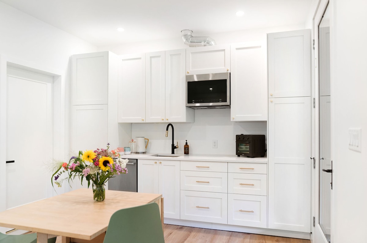 A modern kitchenette is presented with sleek white cabinetry and minimalist design. A small dining table is adorned with a vibrant bouquet of flowers. Essential appliances, including a microwave and refrigerator, are thoughtfully integrated into the space, enhancing functionality.