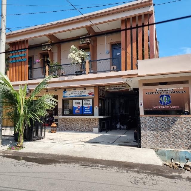A two-story guest house is displayed, featuring a modern façade with wooden accents and a balcony adorned with potted plants. The entrance is welcoming, showcasing signs for amenities and services. A well-maintained pathway leads towards the reception area, with outdoor seating visible on the right.