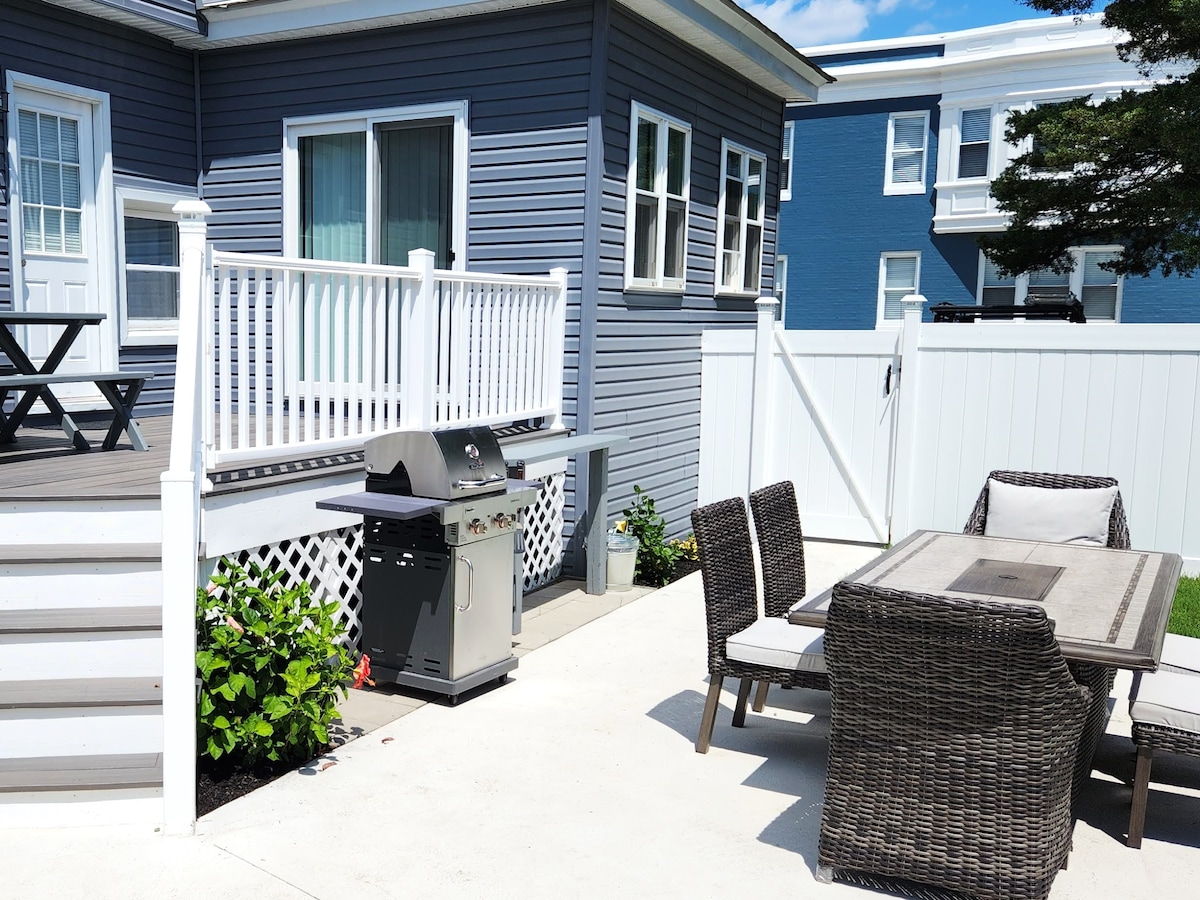 An outdoor deck is seen featuring a dining table surrounded by six chairs. A BBQ grill is positioned nearby, with greenery along the edges. The house is painted in light gray with multiple windows allowing natural light.