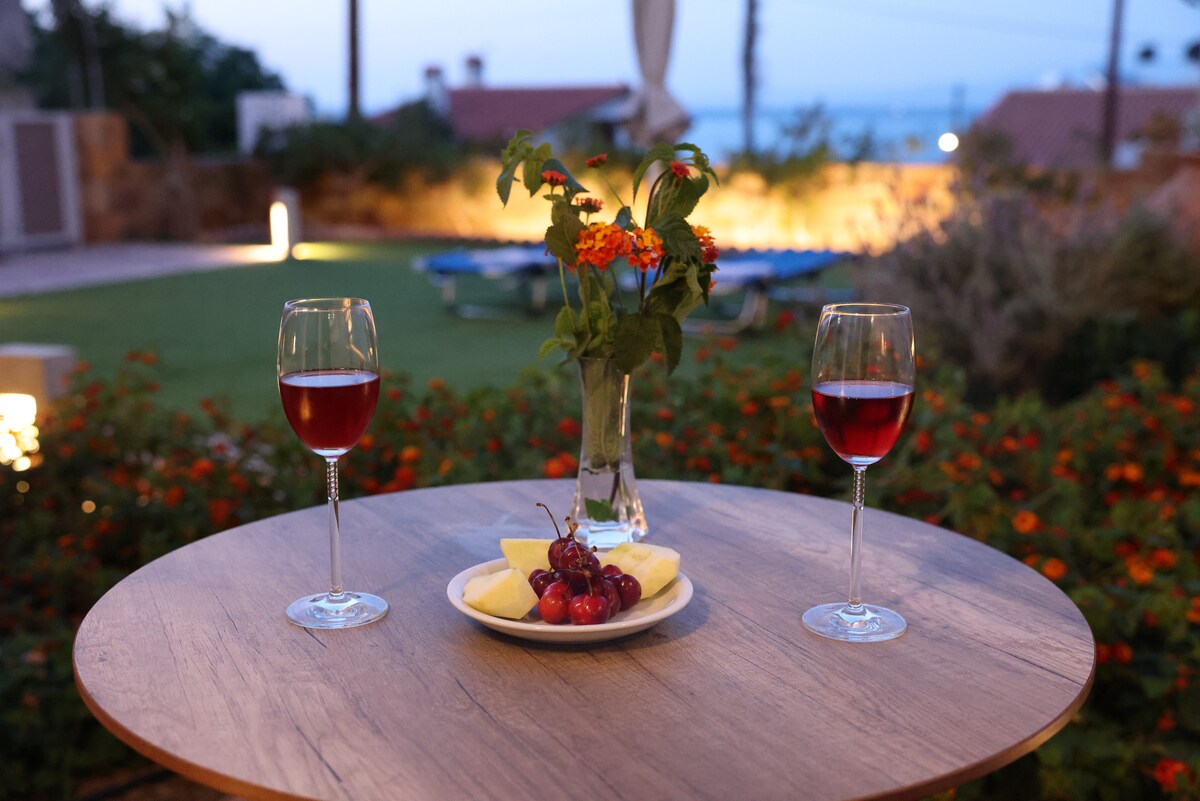 A small wooden table is set with two glasses of red beverage beside a plate of fresh fruit, including grapes and slices of melon. A vase with colorful flowers is centrally placed, while the softly lit outdoor space features a verdant garden in the background.