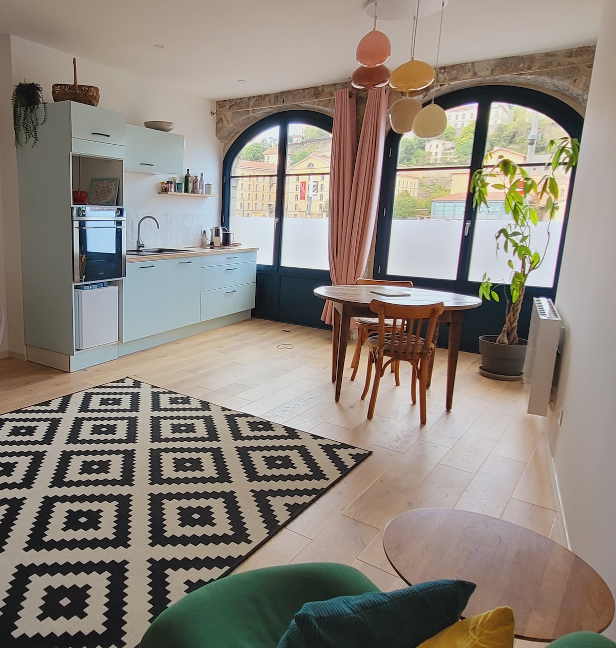 A modern kitchen area is paired with a dining table surrounded by wooden chairs, complemented by a patterned rug on the light hardwood floor. Large windows provide ample natural light, showcasing an indoor plant and decorative lighting that adds character to the space.
