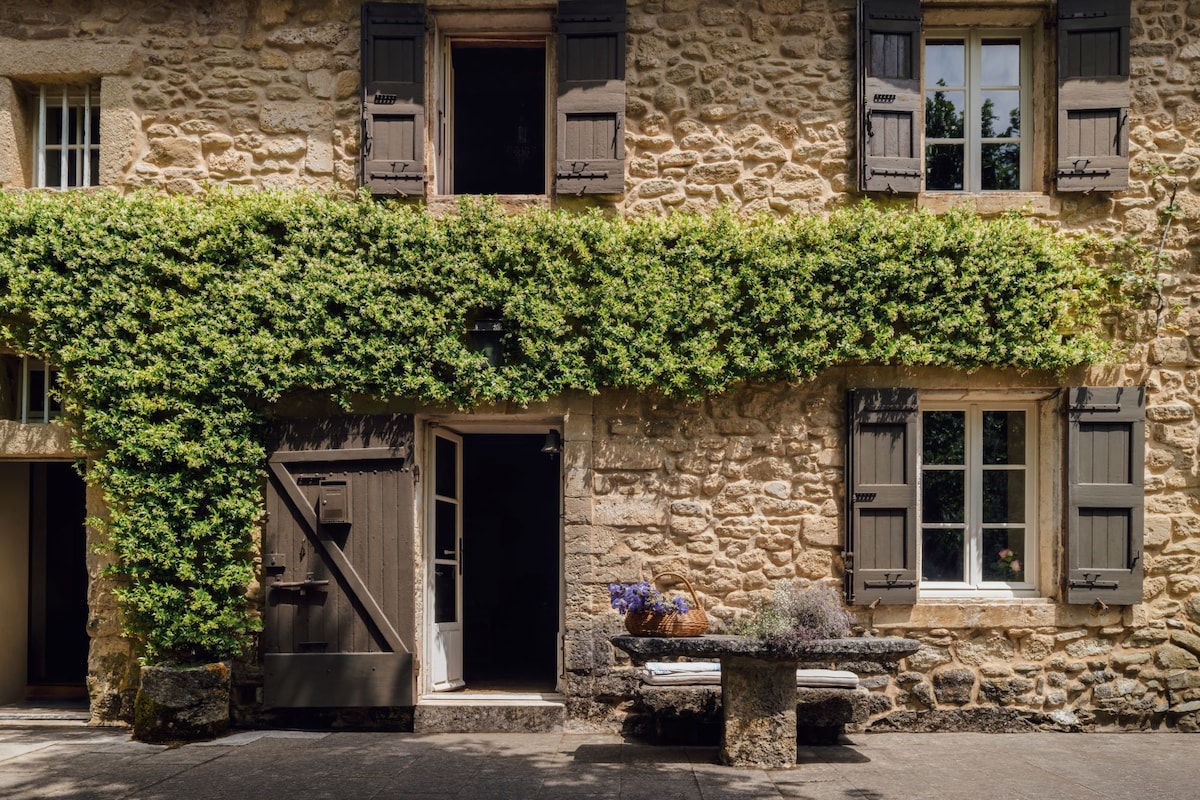 The exterior of a charming farmhouse features rustic stone walls adorned with vibrant green climbing plants. Wooden shutters frame several windows, and a large wooden door is partially open, inviting entry. A stone table, topped with decorative items, enhances the welcoming atmosphere.