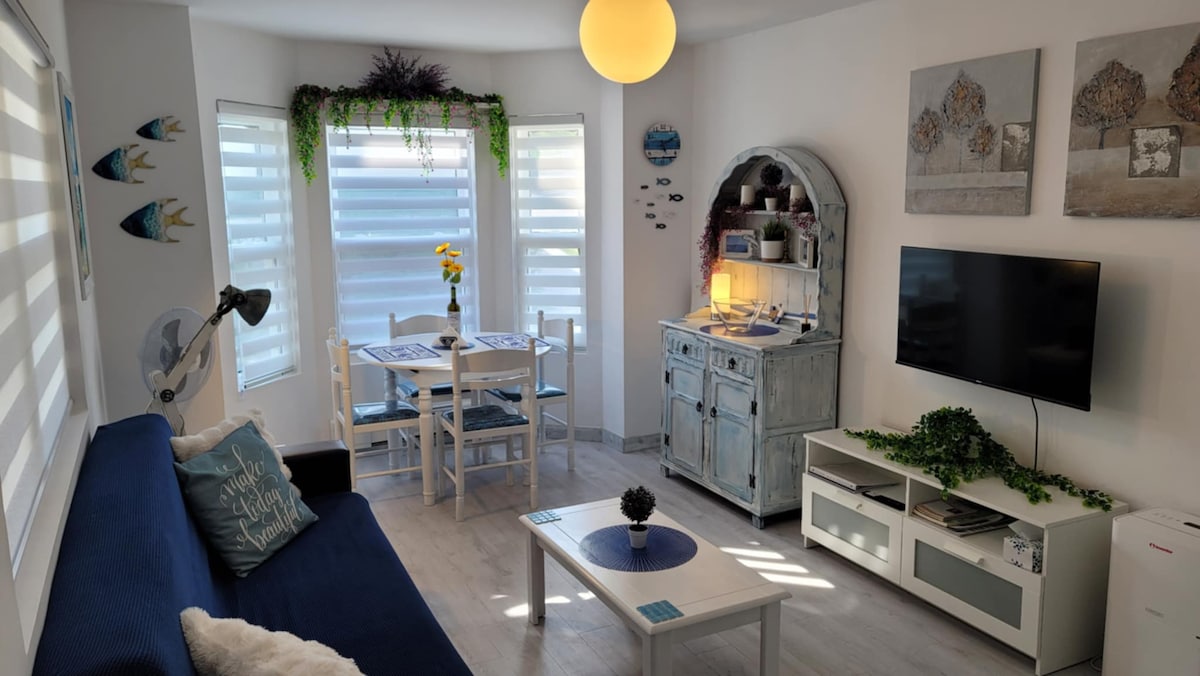 A bright and airy living space features a comfortable blue sofa and a light wooden dining table with four chairs. Natural light filters through the sheer window shades, illuminating decorative wall art and a cozy sideboard display. A television is positioned for easy viewing.