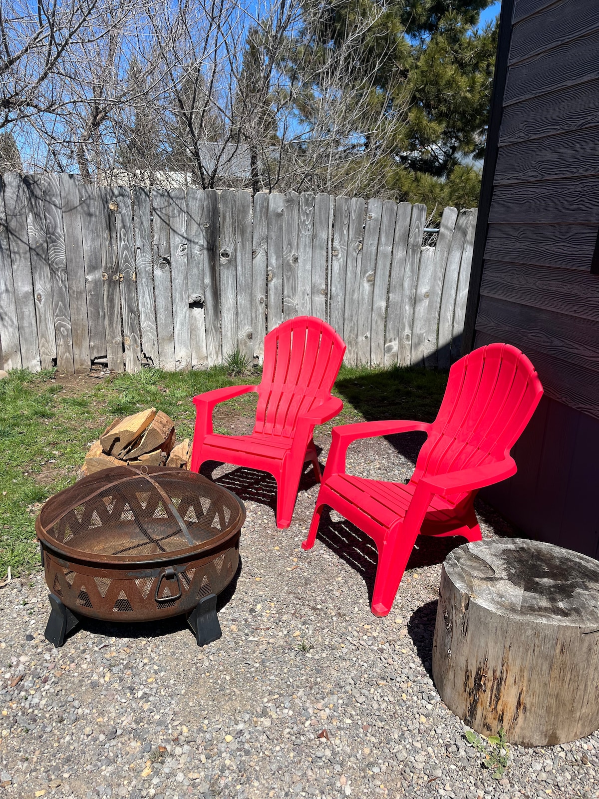 A small outdoor space features two bright red Adirondack chairs positioned beside a round fire pit with a decorative design. A log sits nearby on the gravel surface, and a wooden stump serves as an additional seating option. A wooden fence provides privacy.