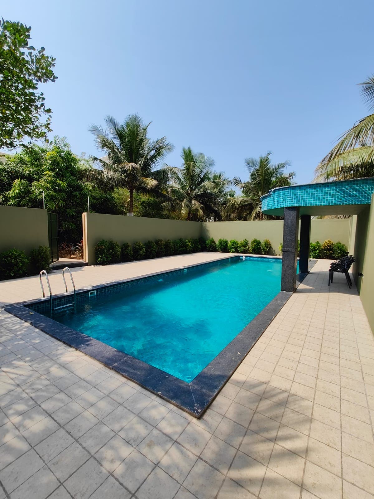A serene swimming pool is positioned within a lush tropical garden. Surrounding palm trees provide shade and enhance privacy. The pool features a tiled deck, with minimal furnishings including a couple of chairs positioned nearby. Clear blue waters reflect the sunlight, creating a calming atmosphere.
