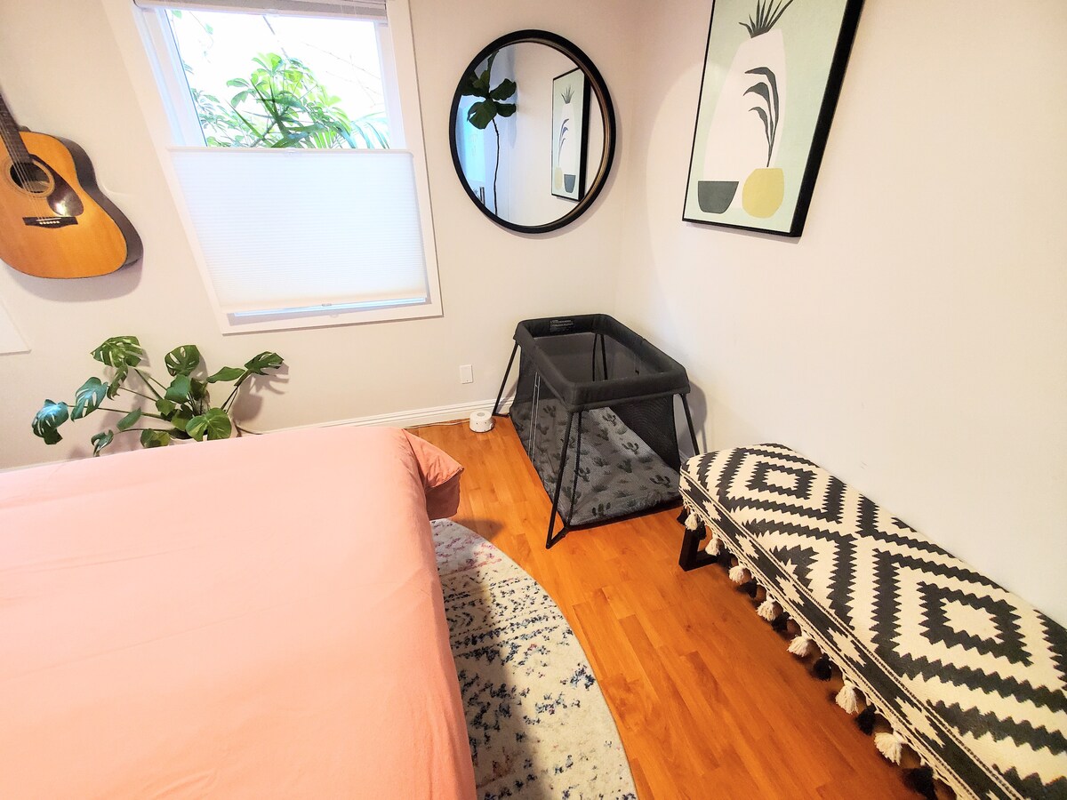 A well-appointed bedroom features a spacious bed covered in soft pink bedding. A large round mirror reflects natural light from a nearby window, enhancing the welcoming ambiance. A black and white patterned bench offers additional seating, while a pack-and-play is positioned nearby for convenience.