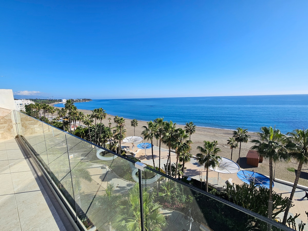 A panoramic view of the shoreline is displayed from a sixth-floor balcony. The scene features a sandy beach bordered by palm trees and a clear, serene sea reflecting the sky. The boulevard below is free of traffic, enhancing the coastal atmosphere.