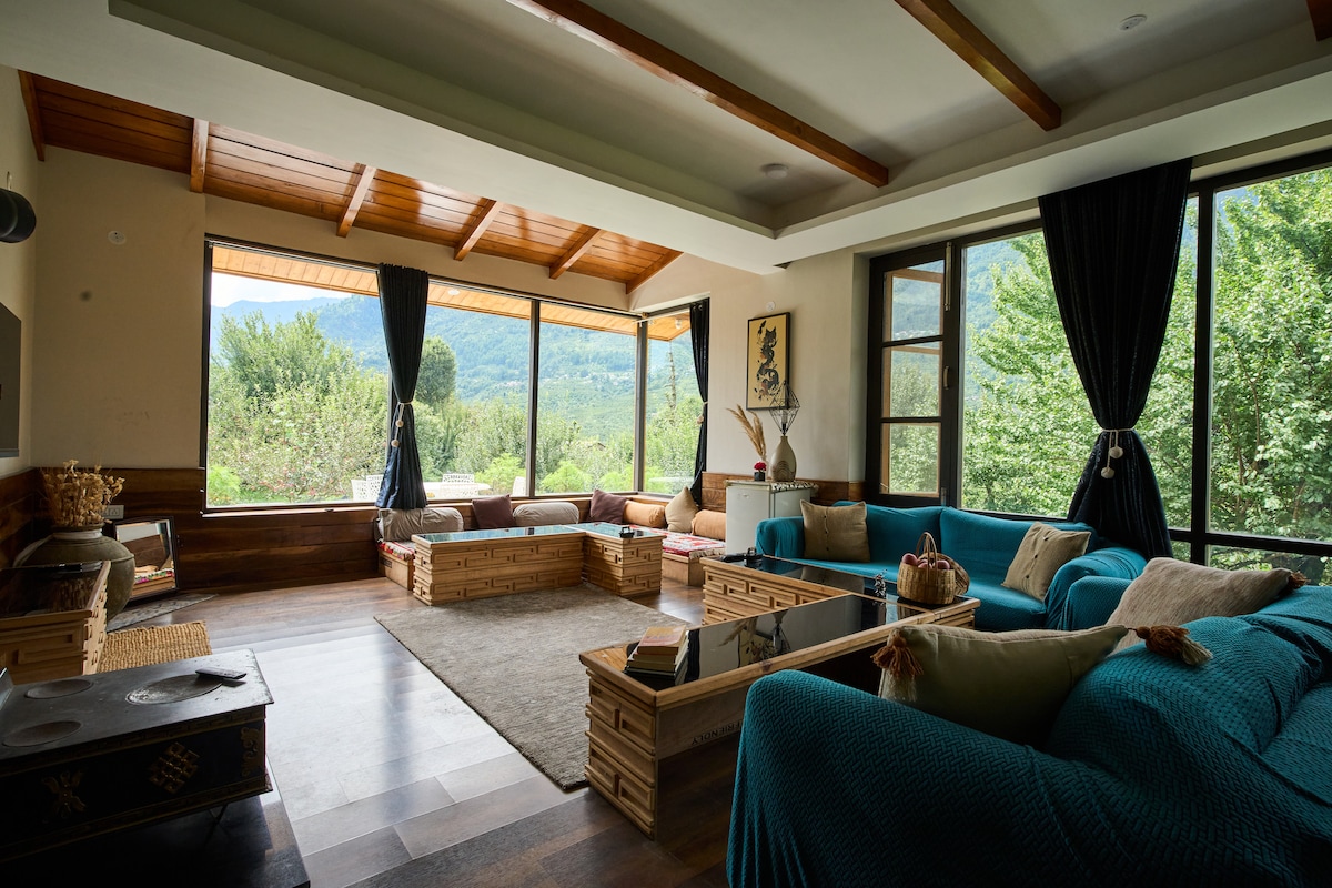 A spacious living area is bathed in natural light from large French windows, offering scenic views of surrounding greenery. Comfortable seating arrangements feature soft, turquoise cushions. The wooden ceiling and floor contribute to a warm ambiance, while a central coffee table adds to the inviting space.