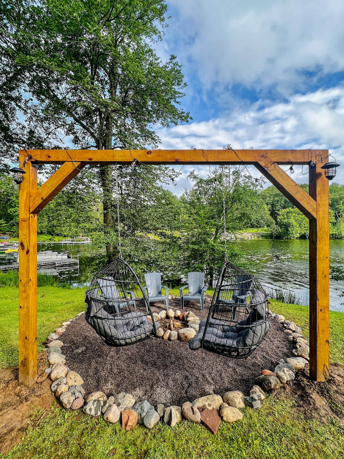 A cozy seating area is framed by a wooden structure, featuring two suspended chairs that gently sway. Surrounded by a circular arrangement of smooth stones, a fire pit offers a gathering space. Lush greenery and the lake create a serene backdrop.