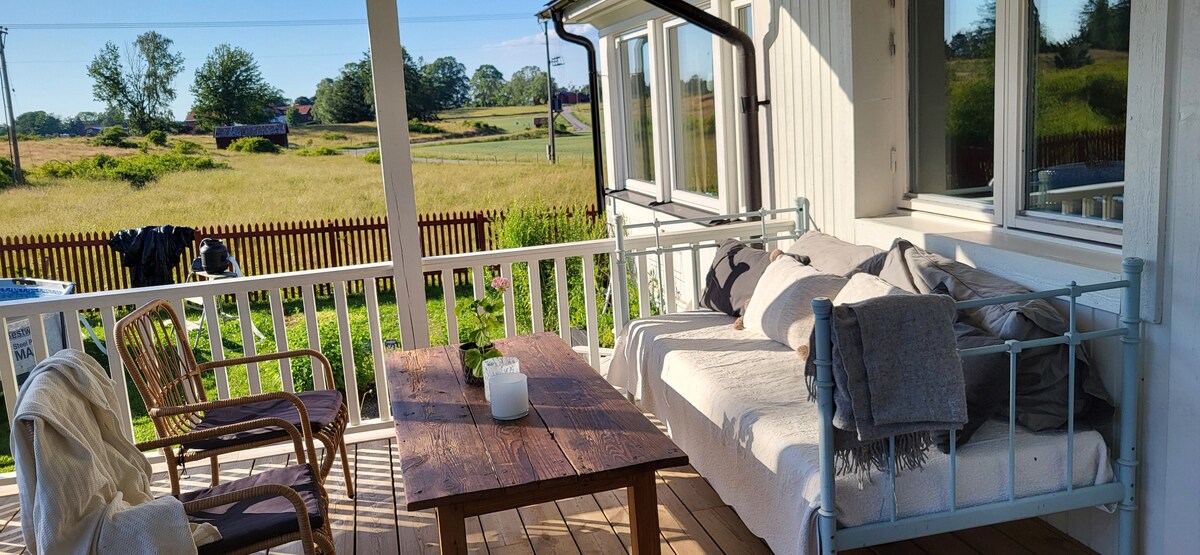 A covered outdoor patio features a wooden table and a comfortable seating area. Surrounding views include open fields and green landscapes. Natural light illuminates the space, creating a relaxed environment for gatherings. Soft blankets and a candle add a welcoming touch.