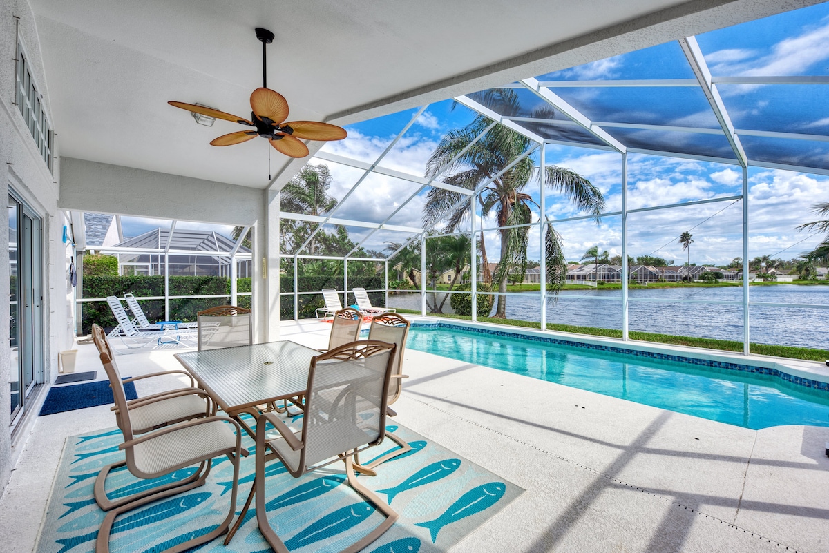 The outdoor lanai features a glass enclosure, providing views of the pool and waterfront. A spacious table with several chairs is positioned on a decorative rug, while lounge chairs are visible nearby. Palm trees frame the scenic landscape under a partly cloudy sky.