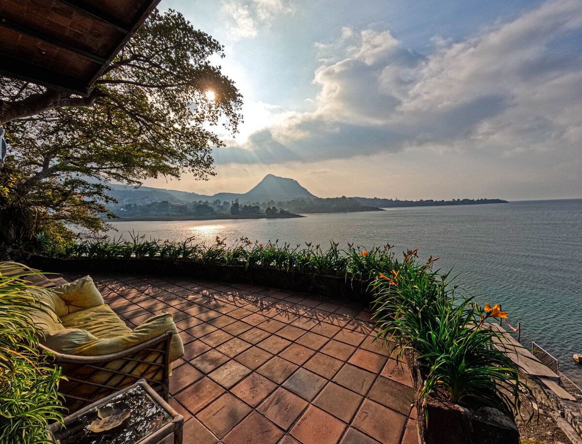 A serene lakeside patio is shown, featuring a comfortable seating area with soft cushions. Surrounding greenery and vibrant flowers frame the space, while the calm lake reflects the soft, natural light filtering through clouds. A distant mountainous landscape further enhances the tranquil setting.