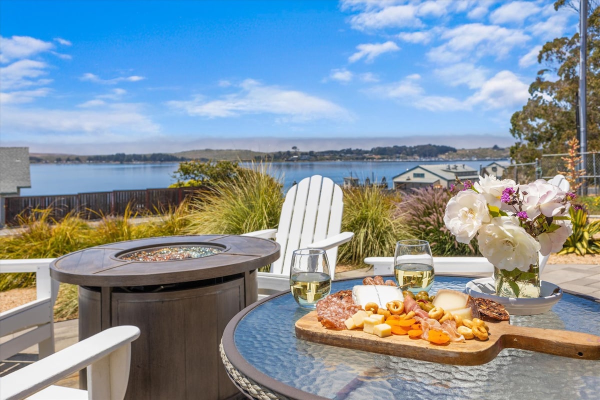An outdoor patio table is set with an assortment of cheeses, meats, and fruits, alongside two glasses of white wine. A fire pit stands nearby, with white adirondack chairs facing a scenic view of calm water and distant trees under a blue sky.