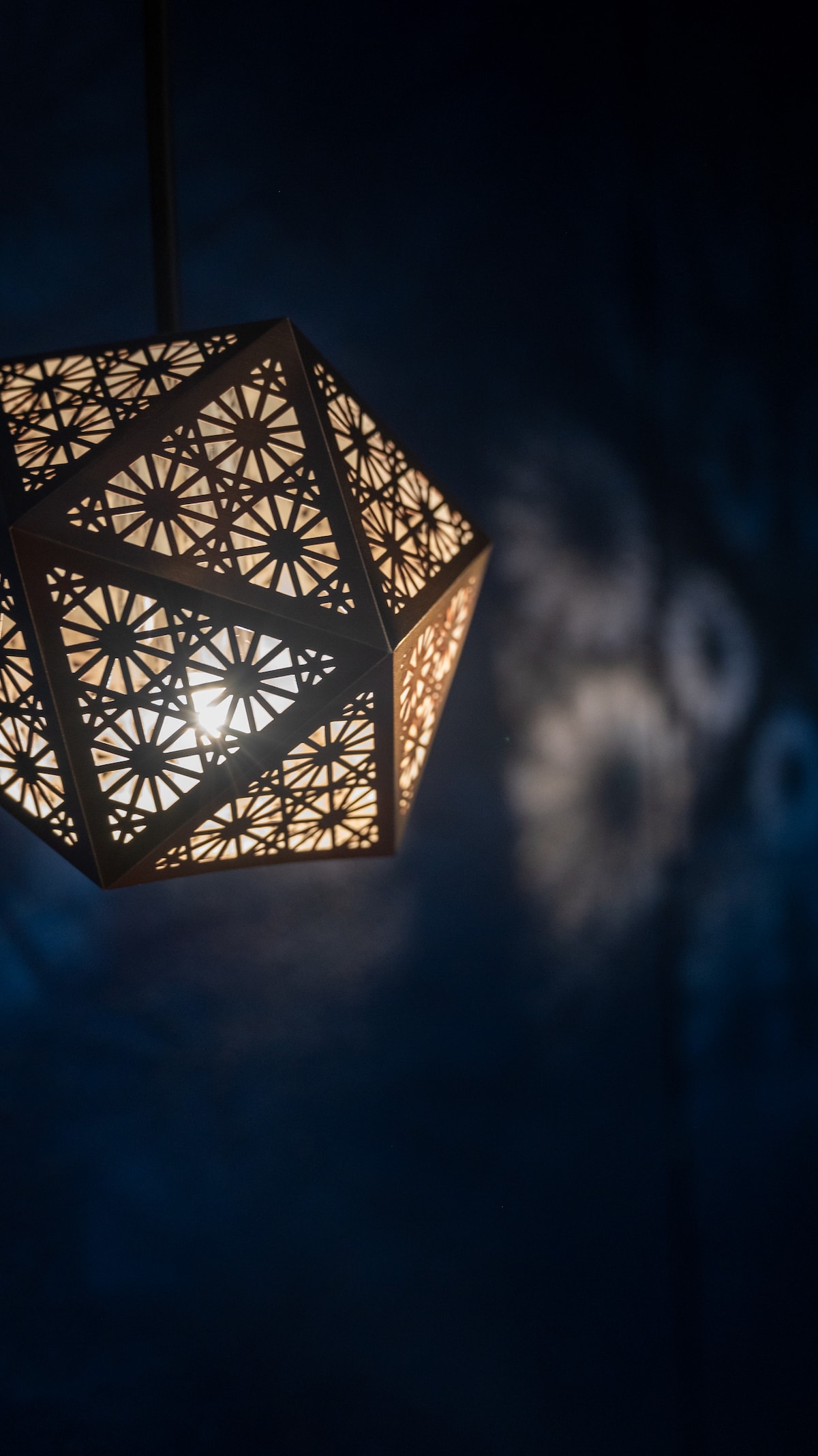 A uniquely designed pendant light is showcased, featuring intricate cut-out patterns that create delicate shadows on the dark wall behind it. The light emits a warm glow, enhancing the ambiance of the space.