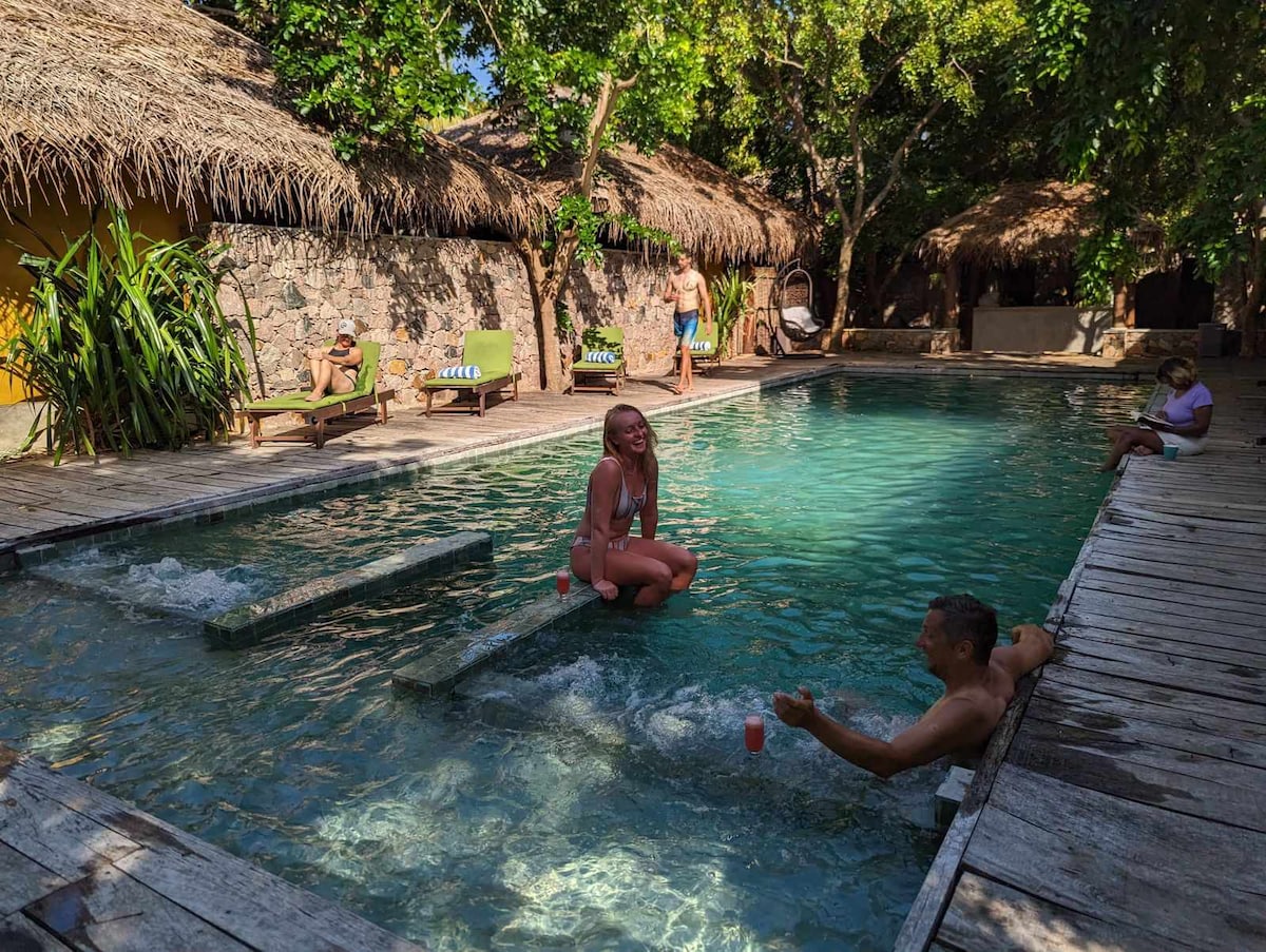 A large swimming pool is surrounded by lush greenery and rustic thatched-roof villas. Guests relax on wooden lounge chairs along the poolside, while another enjoys the water. The calm atmosphere is enhanced by natural light filtering through the trees.