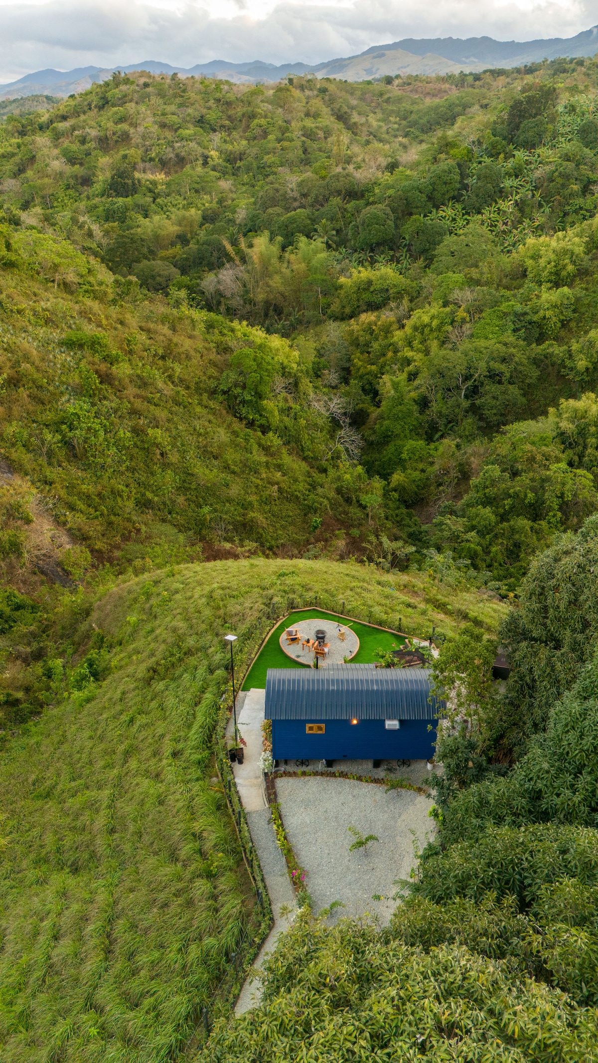 An aerial view captures the Shepherd's Hut surrounded by lush greenery, nestled within a hilly landscape. An outdoor dining area, complete with seating and a small lawn, is visible in the foreground, inviting relaxation amidst nature's tranquility.
