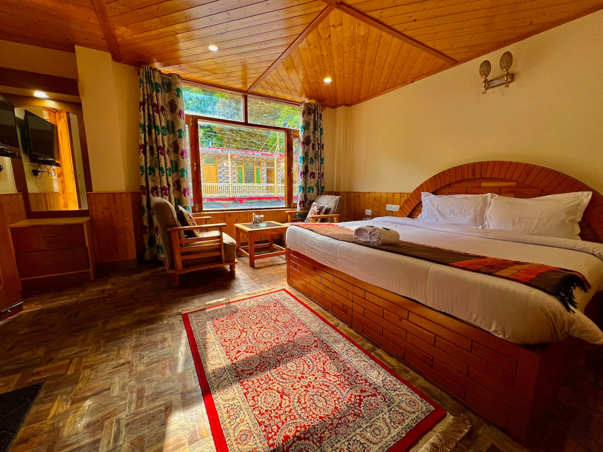 A spacious room features a large bed with a subtle patterned blanket. A wooden ceiling adds warmth, while large windows provide natural light and views. A cozy sitting area includes two chairs and a wooden table. Decorative curtains frame the windows, enhancing the inviting atmosphere.