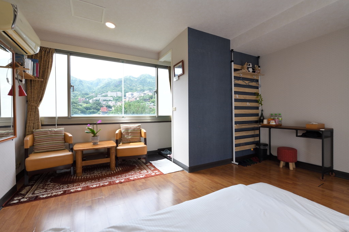 A spacious room features a large window offering a view of the mountains. Two armchairs are arranged around a small wooden table, with decorative items on the table. A desk and chair are positioned against the wall, providing a functional area for work.