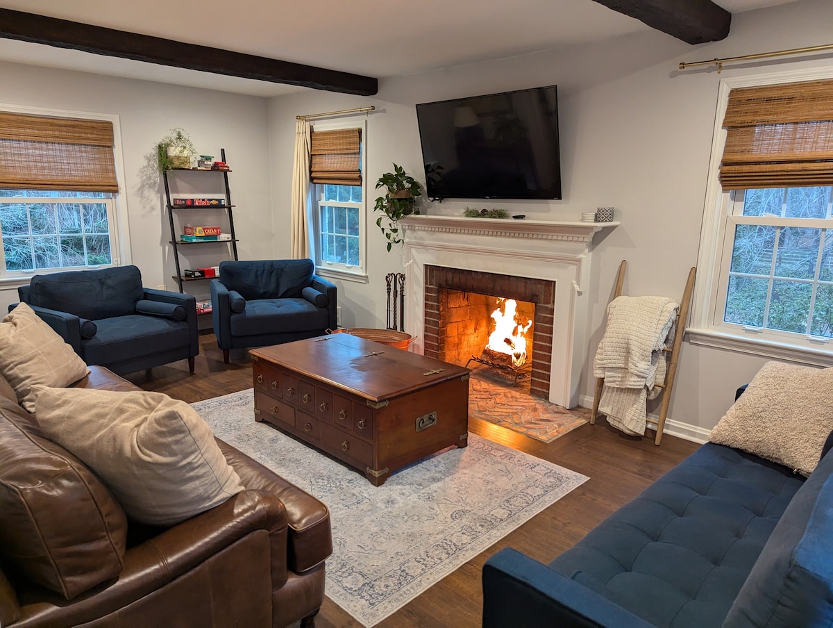 A spacious living room is highlighted by a wood-burning fireplace, with a vibrant flame visible. Comfortable seating includes a leather sofa and plush armchairs around a wooden coffee table. Natural light filters through window shades, enhancing the cozy atmosphere.