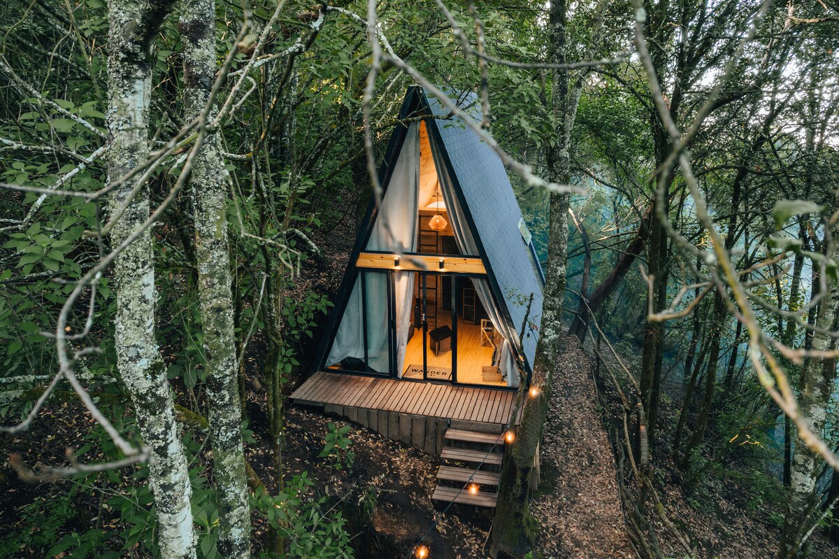 A modern A-frame cabin nestled among trees features a triangular roof and large windows. A wooden deck extends from the entrance, providing outdoor seating. Pathway lights line the approach, offering a welcoming ambiance amid the surrounding forest.
