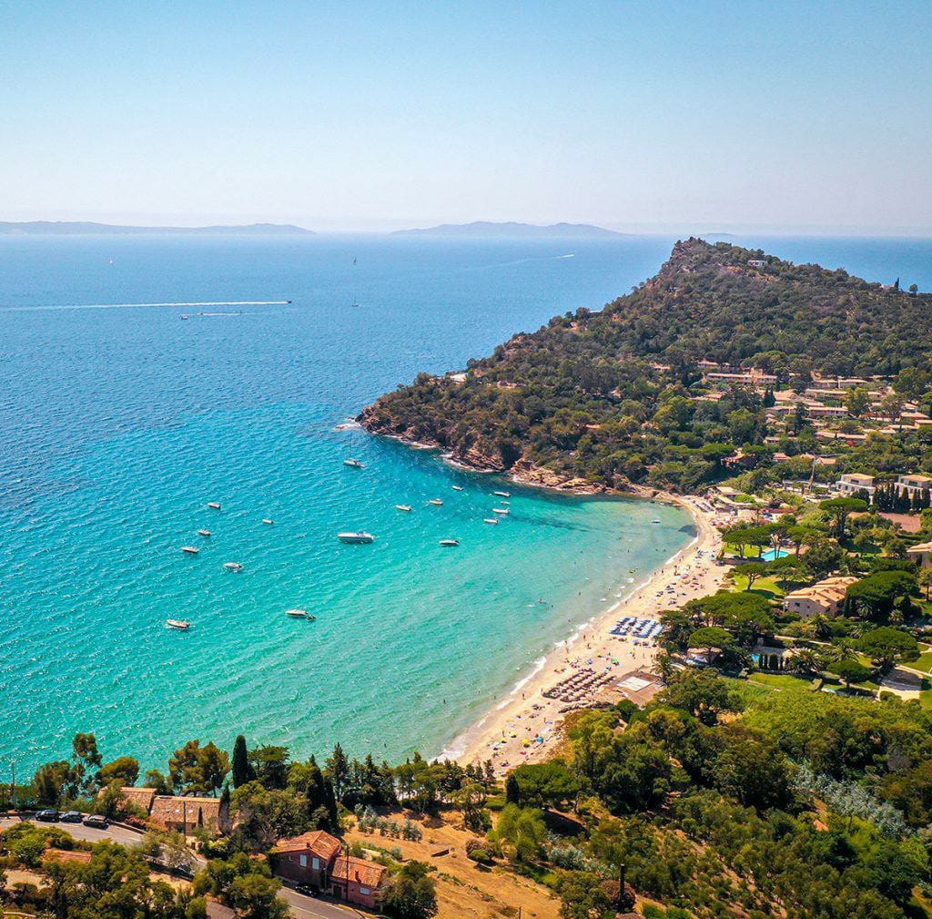 An aerial view captures the coastline, with gentle waves lapping against the sandy beach. Sunbathers enjoy the shore, while boats are seen scattered across the turquoise waters. Lush greenery unfolds along the hillside, complementing the blue Mediterranean landscape.