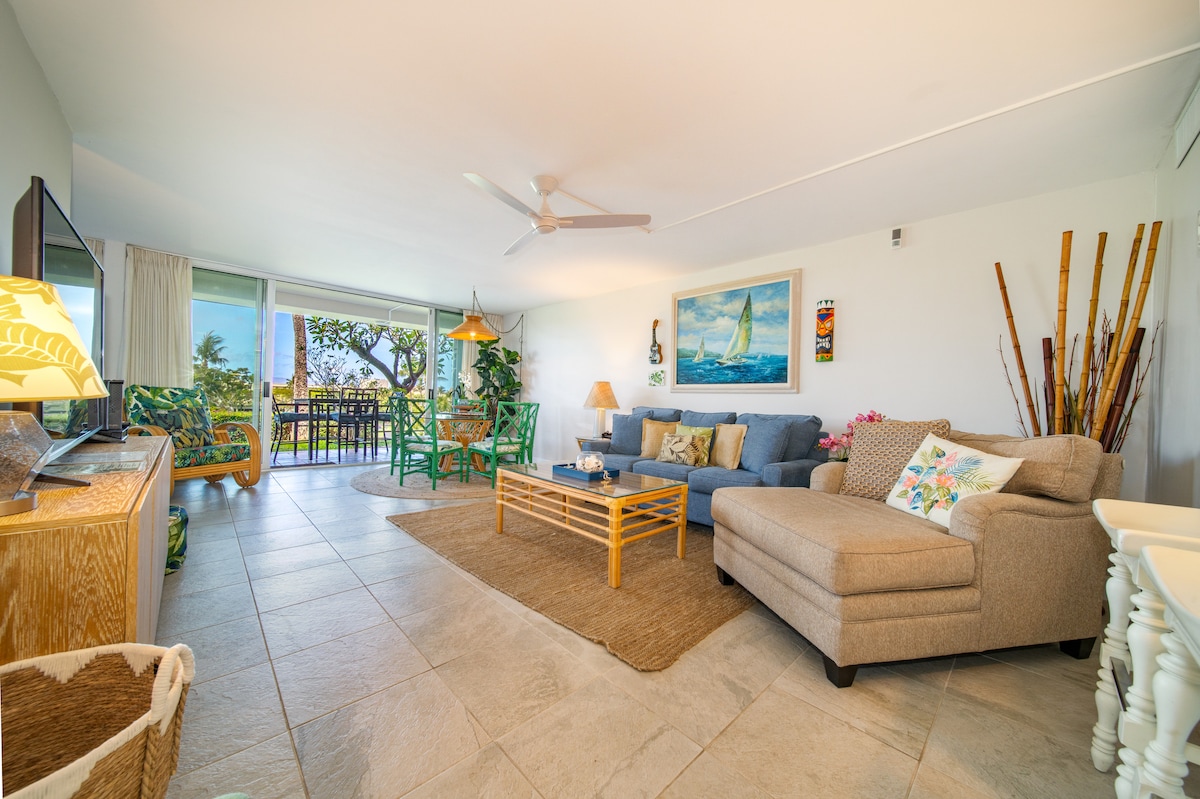 A spacious living area features a light-coloured sectional sofa, a coffee table, and a flat-screen TV. Large sliding glass doors provide access to the lanai, allowing natural light to fill the space. Decor includes tropical artwork and a mix of comfortable seating.