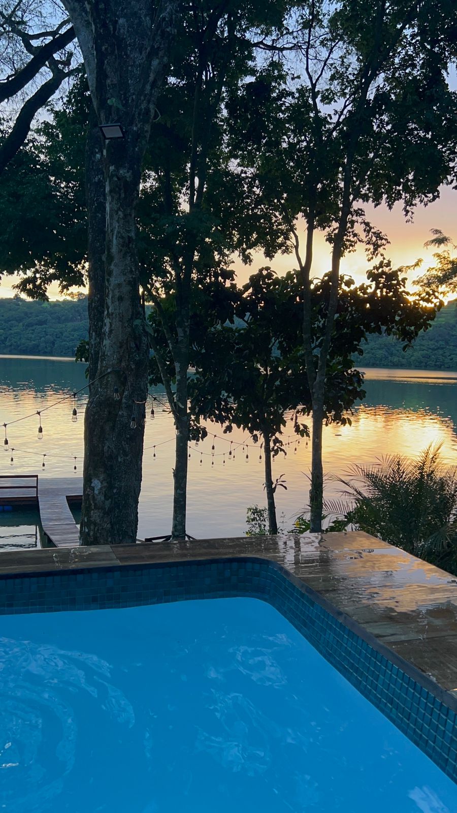 A tranquil view of a private pool surrounded by trees, with the lake reflecting the soft colors of a sunset. The water appears calm, enhancing the serene atmosphere of the outdoor space.
