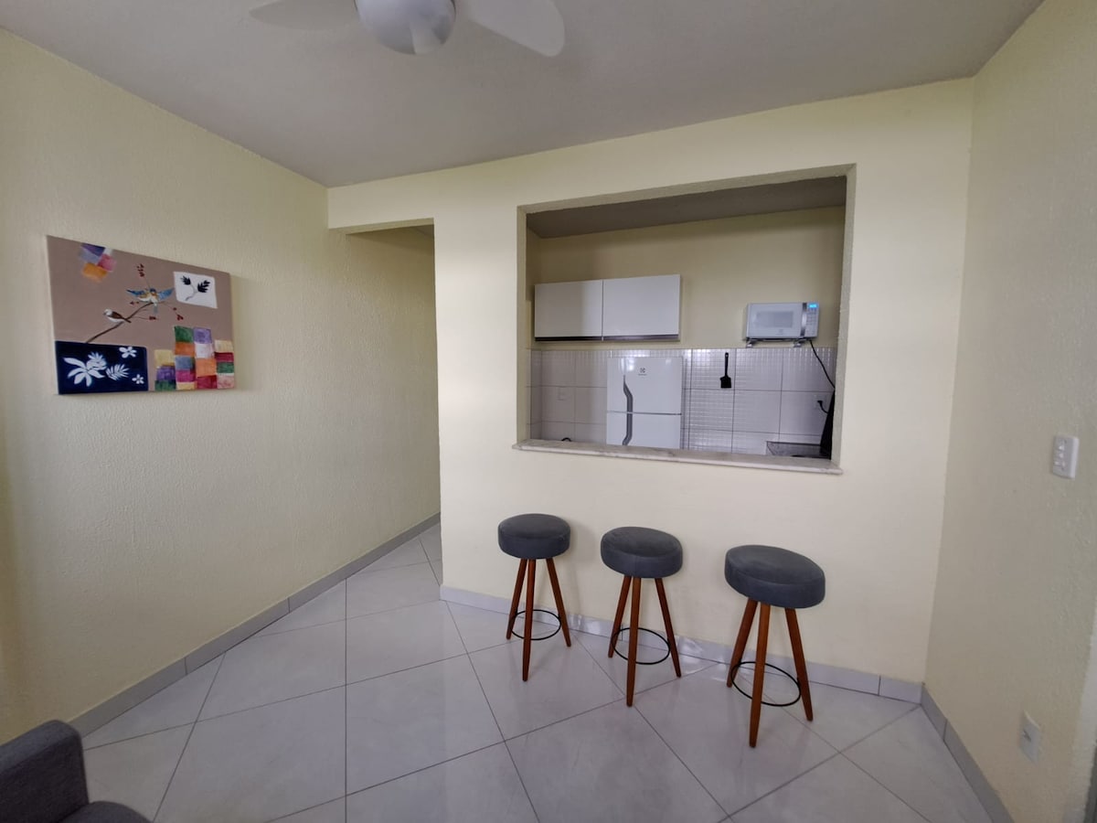 A small dining area features three wooden stools positioned in front of a wall-mounted kitchen counter. The space includes a modern kitchen with essential appliances visible in the background, complemented by a vibrant piece of art on the wall, adding a touch of color to the light-colored room.