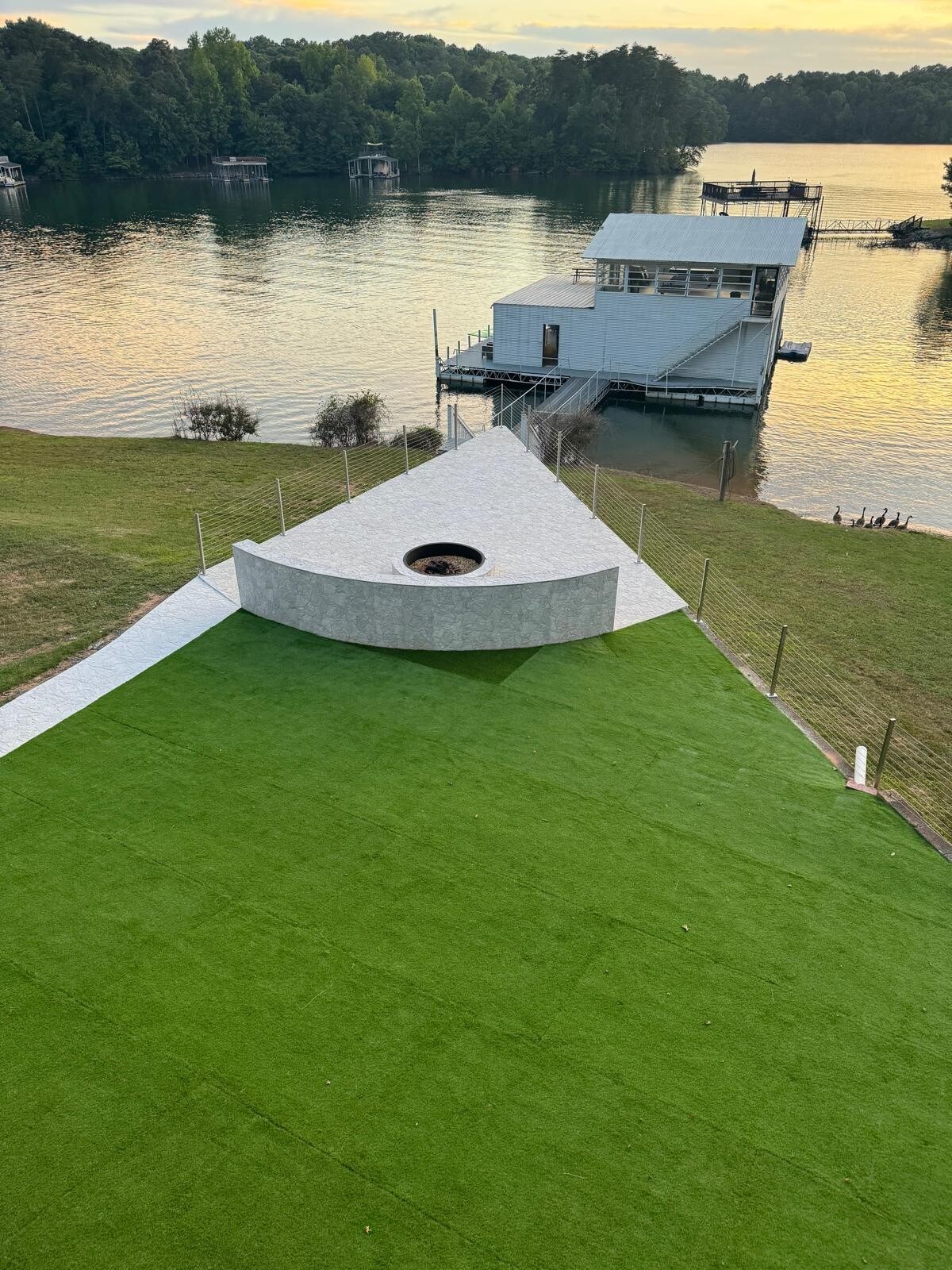 A landscaped yard with synthetic grass leads to a modern triangular fire pit. The backdrop features a tranquil lake and a boat dock, enhancing the serene lakeside setting. The area is surrounded by gentle slopes and trees, complementing the peaceful ambiance.
