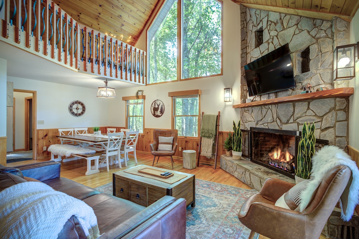 The great room features a stone fireplace with a warm fire, surrounded by comfortable seating. Large windows allow natural light to flood in, highlighting the wooden ceilings and beams. A dining area, visible in the background, is furnished with a table and white chairs.