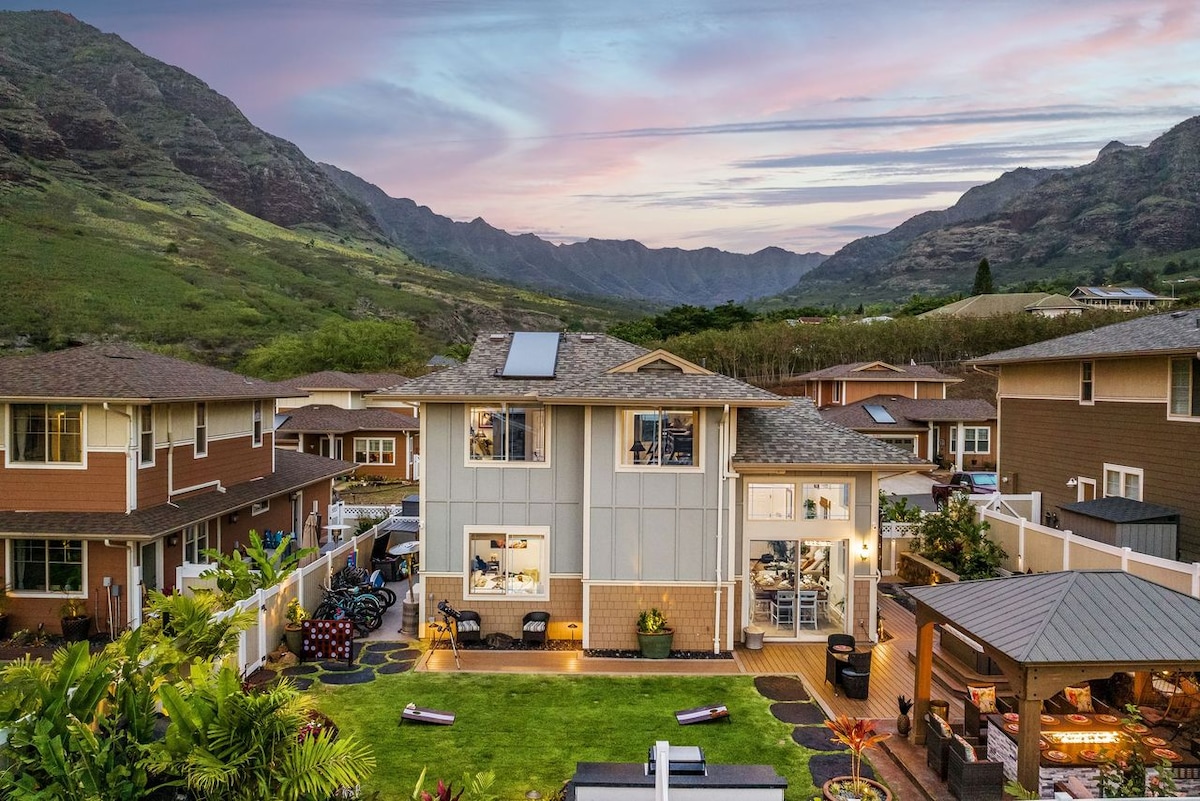 The two-story residence is surrounded by lush greenery, with the backdrop of the Wai'anae Mountain Range. Outdoor spaces feature a manicured lawn and a gazebo area, making it ideal for relaxation. A serene sky during twilight casts a warm glow over the neighborhood.