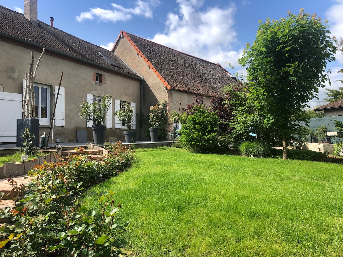 A serene garden area is presented with well-maintained green grass and various plants in large pots. Stone pathways lead through the space, with a backdrop of neighboring structures featuring traditional roofs. The scene is enhanced by a clear blue sky and scattered clouds.