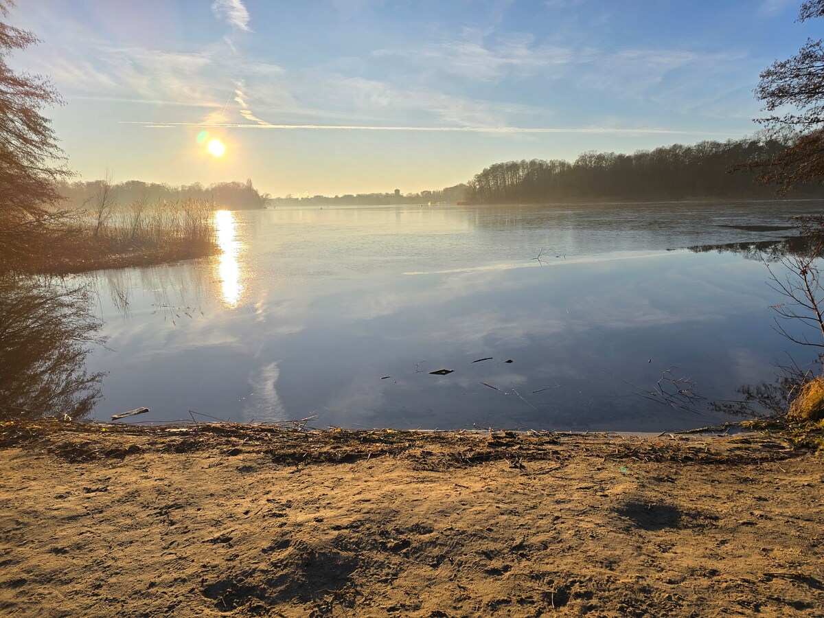 The image captures a serene lakeside view at sunrise, with the sun's rays reflecting on the calm water's surface. Gentle ripples are visible, surrounded by trees on the far shore. The sandy foreground transitions to the water's edge, creating a tranquil atmosphere.