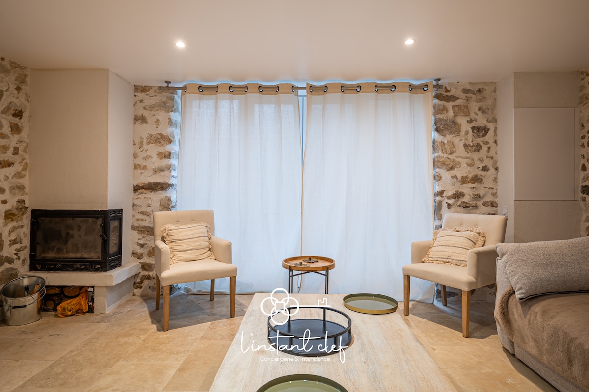A cozy living area features two light armchairs positioned opposite a modern fireplace. Natural light flows through sheer curtains, while the stone walls add a rustic charm. A low table with circular trays sits in the center, inviting relaxation in this tranquil space.