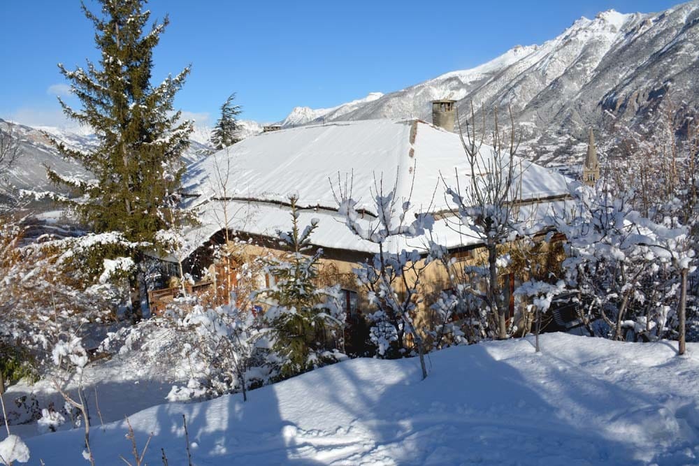 A charming mountain house is partially covered with snow, surrounded by trees and a serene winter landscape. Snow blankets the ground, highlighting the natural beauty of the area. Majestic mountains rise in the distance under a clear blue sky.