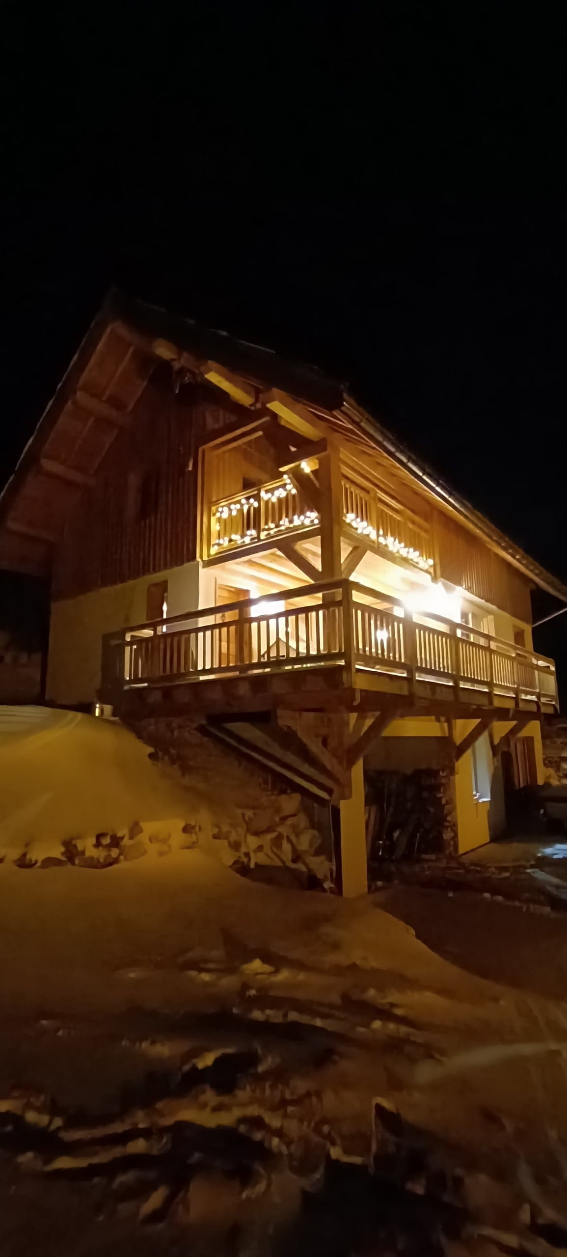 The chalet is illuminated at night, highlighting its wooden structure and balcony. Snow blankets the ground, and the outdoor lighting creates a warm glow against the dark backdrop of the evening sky.