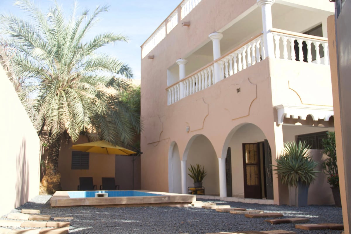 The outdoor area features a private pool surrounded by a wooden border, with two lounge chairs positioned nearby. A yellow umbrella provides shade. The villa showcases a two-story structure with arched doorways and decorative balconies, framed by palm and ornamental plants.