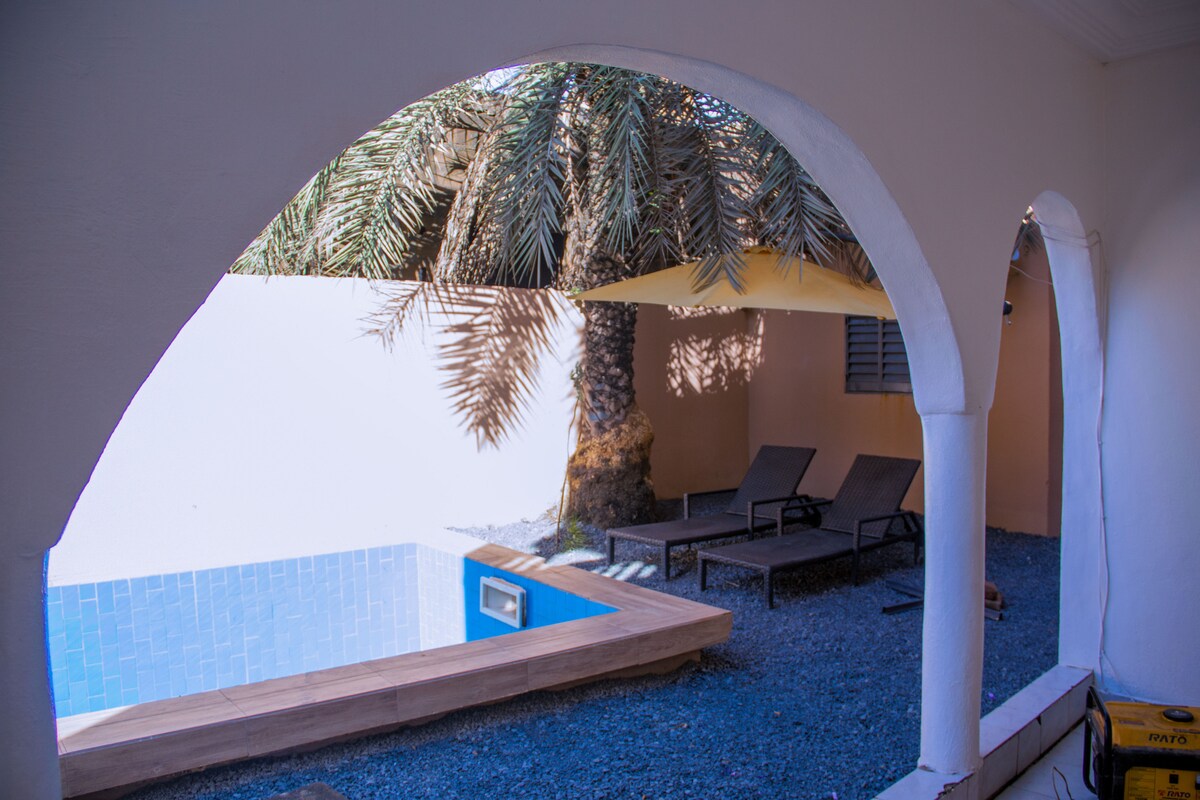 The outdoor area features a private swimming pool surrounded by palm trees. A shaded terrace with lounge chairs provides a place to relax. Arched architectural details frame the view, enhancing the tranquil setting.