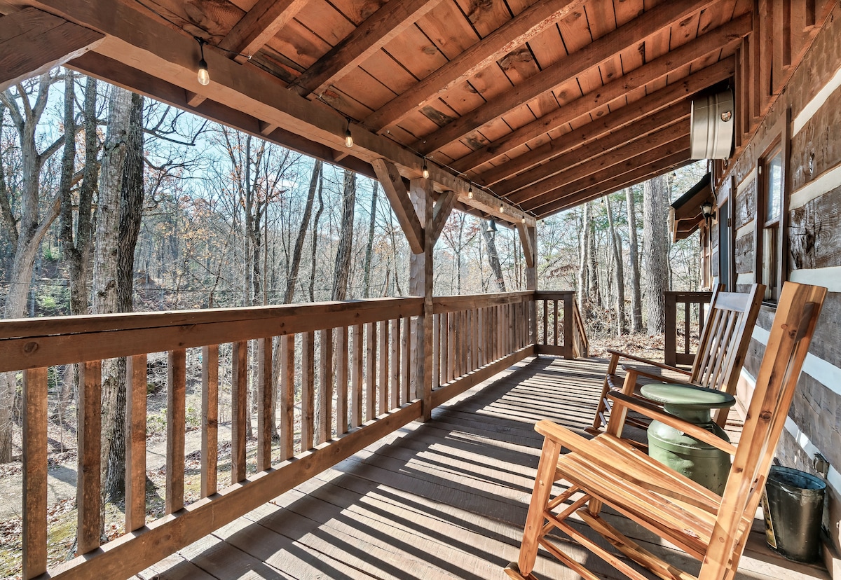 A covered porch with wooden beams features two rocking chairs positioned near the railing. Natural light filters through the trees, creating a serene outdoor space. The porch overlooks the surrounding wooded area, inviting relaxation in a peaceful setting.